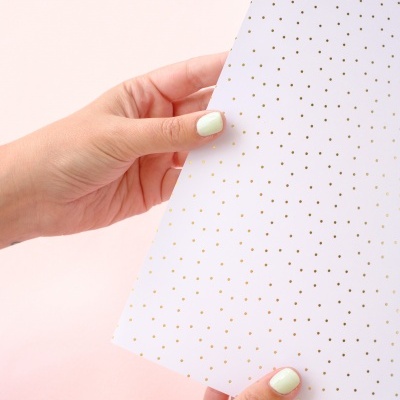 Folha branca com pontos dourados segurada por mãos com unhas claras, fundo rosa