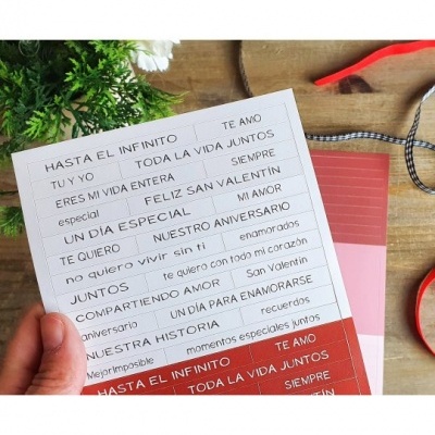 Folha com frases em castelhano para cartão, mão segurando, fundo de madeira, plantas e fitas ao redor