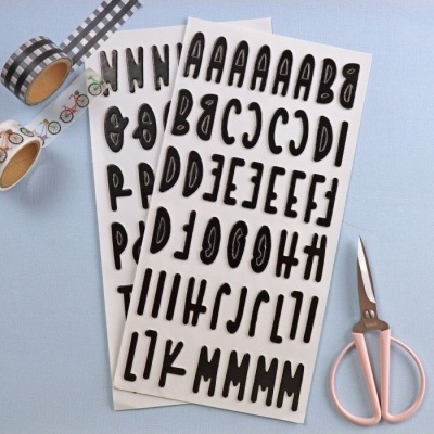 Folhas com letras autocolantes negras, rolos de fita adesiva decorativa de bicicletas e xadrez, e tesoura rosa em fundo azul.