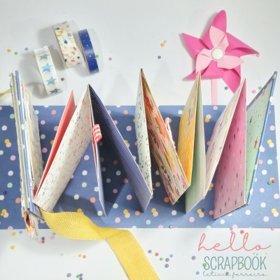 Livro de scrapbook com várias capas coloridas e padrões, rolos de fita decorativa, catavento rosa e texto hello SCRAPBOOK