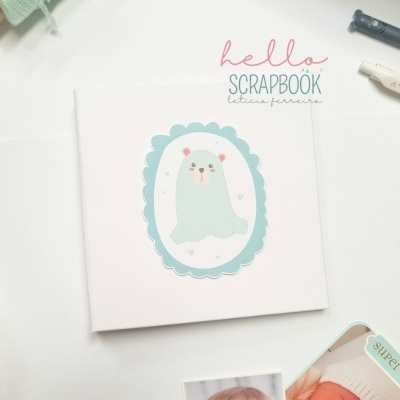 Álbum com ilustração de foca azul claro e borda scalloped azul sobre mesa branca com fotos de bebés.