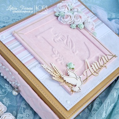 Livro decorativo cor-de-rosa com relevo e adornos florais e em madeira com palavra Amém sobre fundo rendado azul.