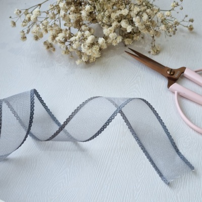 Fita cinza decorativa em organza com bordas bordadas, flores secas e tesoura rosa numa superfície branca.