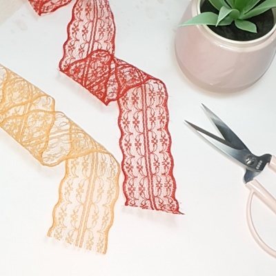 Tiras de renda vermelha e amarela, tesoura rosa e vaso com planta verde sobre fundo branco.