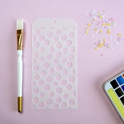 Pincel, stencil, lantejoulas e paleta de aquarela sobre fundo rosa.