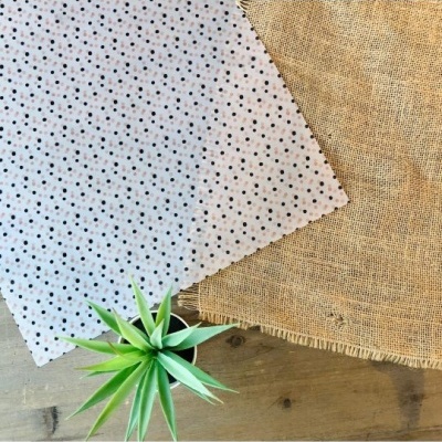 Dois tecidos, um branco com pontos pretos e um castanho rústico, sobre mesa de madeira com planta verde.