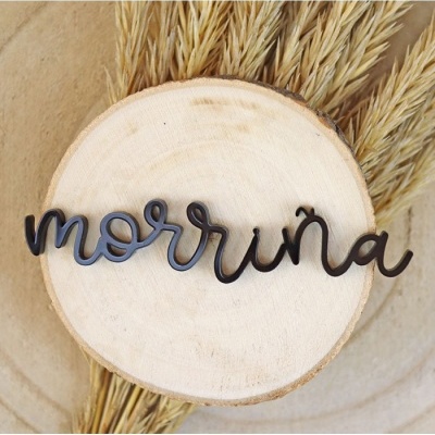 Produto decorativo em madeira preta com a palavra 'morriña' sobre base circular de madeira clara e plantas secas ao fundo.