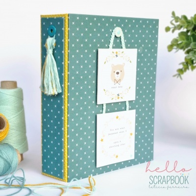 Álbum scrapbook verde-azulado com padrão de cruzes brancas e ilustrações de urso e texto penduradas na capa