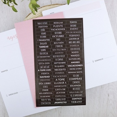 Folha preta com texto branco com várias palavras em espanhol sobre calendário, colocada em cartão rosa e agenda em fundo de madeira clara