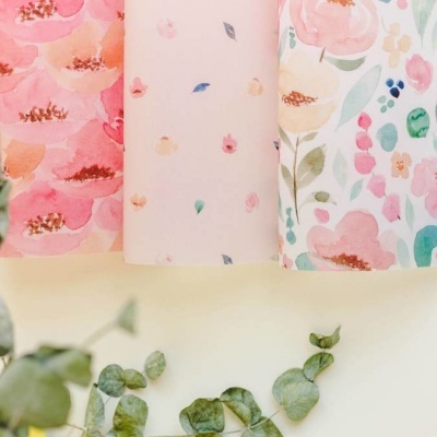 Rolos de papel com padrões florais coloridos em tons rosa e verde, sobre superfície clara com folhas verdes