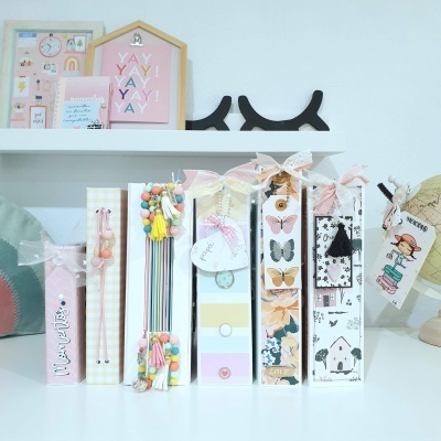 Sete cadernos decorativos coloridos com laços e acessórios sobre mesa branca, prateleira com quadros ao fundo