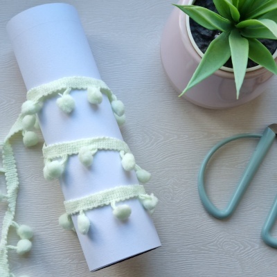 Rolo branco com fita de tecido creme com pompons ao lado de vaso rosa com planta e ferramenta azul