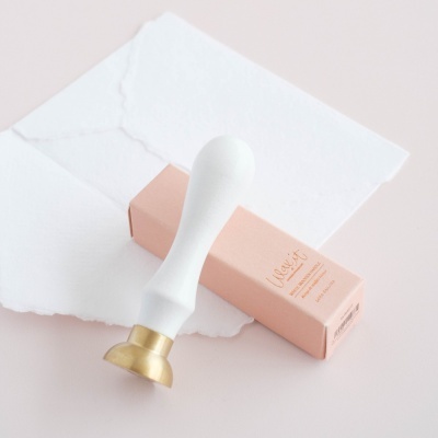 Selo de cera com cabo branco e cabeça dourada e caixa cor de rosa sobre mesa com papéis brancos