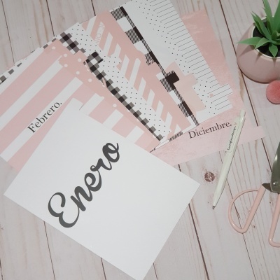 Folhas de calendário com os meses Janeiro, Fevereiro e Dezembro em rosa, branco e preto, com tesoura e lápis sobre superfície clara de madeira