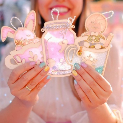 Ornamentos de madeira decorativos com temas festivos e glitter segurados por mãos femininas.