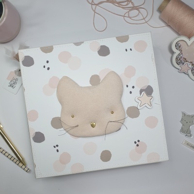Caderno decorado com aplique de gato em tecido e bolinhas pastel, caneta dourada, fio e enfeites de estrelas numa mesa branca