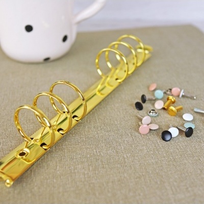 Anel dourado de argolas para fichário com botões coloridos e caneca branca ao fundo.