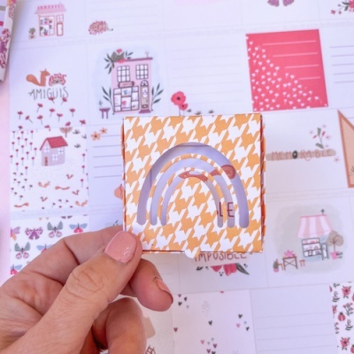 Mão segurando adesivo de arco-íris lilás em caixa com padrão laranja, fundo de cartões ilustrados