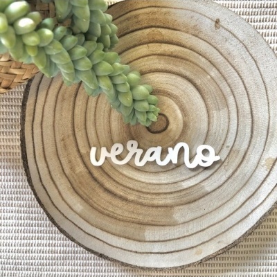 Palavra decorativa 'verano' em madeira branca sobre base circular com textura de madeira e folhas verdes próximas.