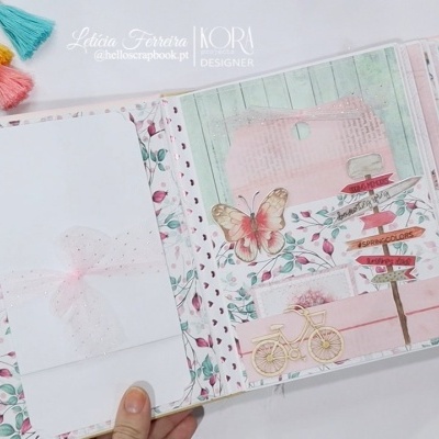 Álbum de scrapbook decorado com padrões florais, bicicleta amarela, borboleta, marcador colorido e envelope branco com laço.