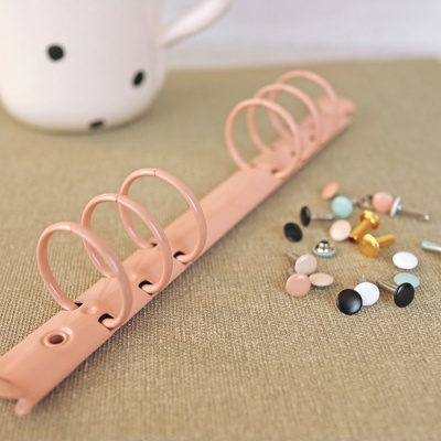 Anel para fichário rosa com argolas e rebites coloridos sobre tecido bege.