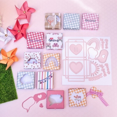 Caixas decorativas pequenas coloridas com padrões e decorações em papel sobre fundo rosa com flores de papel.