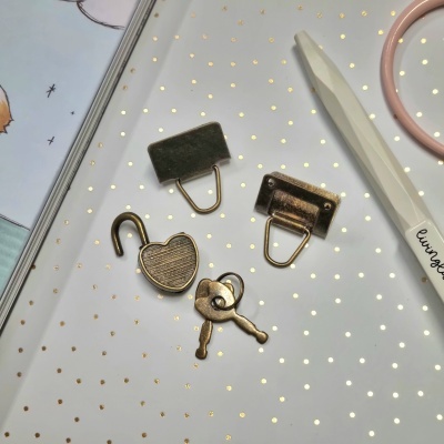 Quatro objetos metálicos bronze, cadeado coração aberto com duas chaves, dois suportes, caneta branca living, livro e fundo branco com pontos dourados.