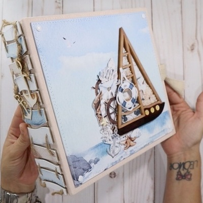 Álbum scrapbook com capa azul e veleiro de madeira, conchas e cordas, mãos segurando
