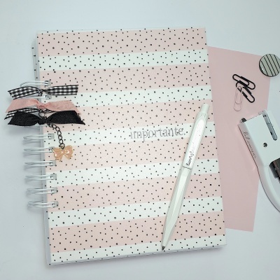 Caderno espiral riscado rosa e branco com pontos pretos e três laços decorativos, caneta branca e acessórios de escritório sobre mesa branca