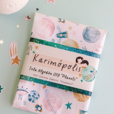 Tecido de algodão estampado com planetas e estrelas em embalagem sobre mesa azul clara com decoração de estrelas e globo lunar.
