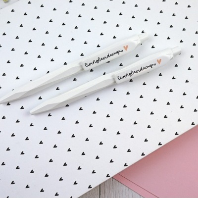 Dois esferográficos brancos com escrita e coração laranja sobre papel com corações pretos.