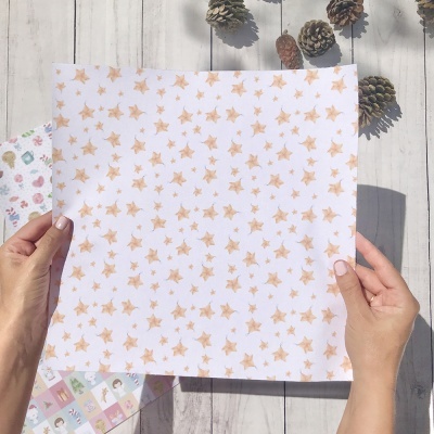 Pano branco com estrelas cor de rosa e laranja segurado por duas mãos sobre mesa de madeira clara com pinhas e papéis coloridos