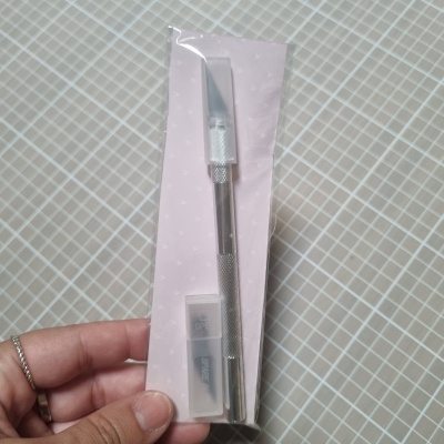 Instrumento metálico para corte com protetor plástico numa embalagem rosa com padrão de corações brancos