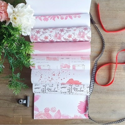 Rolos de papel decorativo com estampas florais em rosa e branco, fitas e flores sobre mesa de madeira.