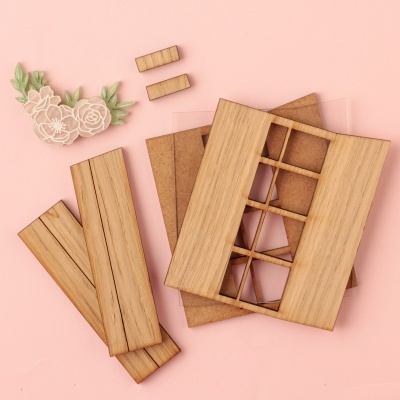 Peças de madeira clara desmontadas com aplicação decorativa de flores brancas e folhas verdes sobre fundo rosa