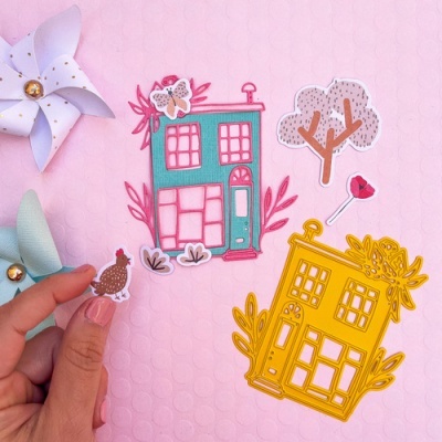 Autocolantes decorativos coloridos com casas, árvore, flor e galinha num fundo rosa texturizado.