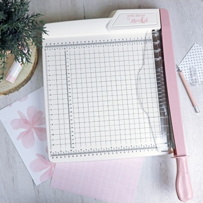 Guilhotina de papel branca e rosa com grelha quadriculada ao lado de folhas e uma planta