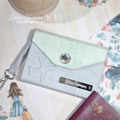 Carteira decorativa verde água e cinza com botão brilhante e etiqueta Letícia Ferreira junto a passaporte e ilustração.