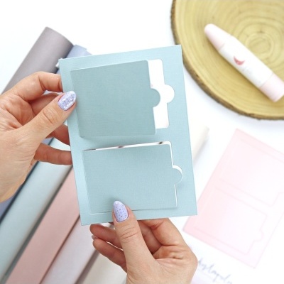 Cartão azul claro com recortes para papel segurado por mãos femininas com unhas pintadas lilás, em mesa com cilindros de papel e batom rosa.