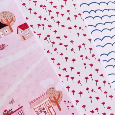 Três tecidos com padrões diferentes lado a lado, um rosa com casinhas e árvores, outro branco com flores vermelhas pequenas, e o terceiro branco com ondulações azuis.