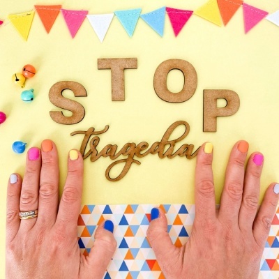 Palavras 'STOP Tragedia' em madeira e metal com mãos de unhas coloridas sobre fundo amarelo com bandeirinhas e pequenos sinos coloridos