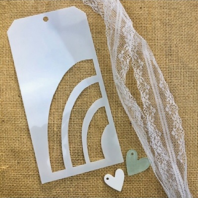 Stencil plástico branco com padrão arco-íris sobre juta com fita de renda e corações decorativos.