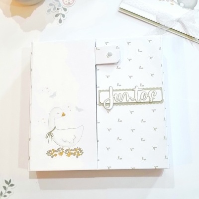 Caderno ou álbum branco com padrão cinzento e ilustração de ganso e texto Juntos, com recortes decorativos e flor branca em vaso