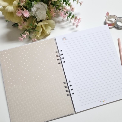 Caderno aberto com capa interna bege com padrões brancos e páginas pautadas, com flores e material de escrita ao lado