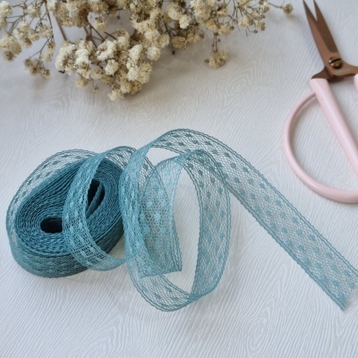 Fita decorativa azul em malha, flores secas e tesouras cor-de-rosa sobre superfície clara.