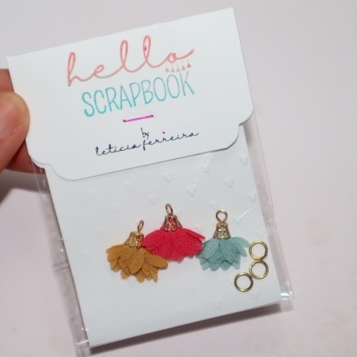 Pacote de scrapbooking com mini pompons coloridos e argolas em embalagem branca com texto.