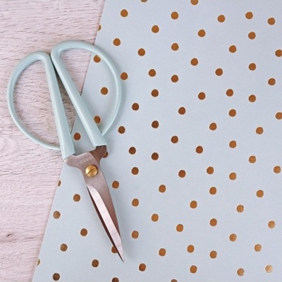 Tesoura com cabo azul claro e papel branco com pontos dourados sobre madeira clara