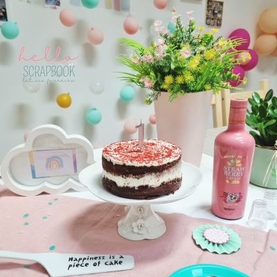 Bolo de aniversário sobre prato branco com vela 1, vasos com plantas, garrafa rosa e decoração festiva colorida