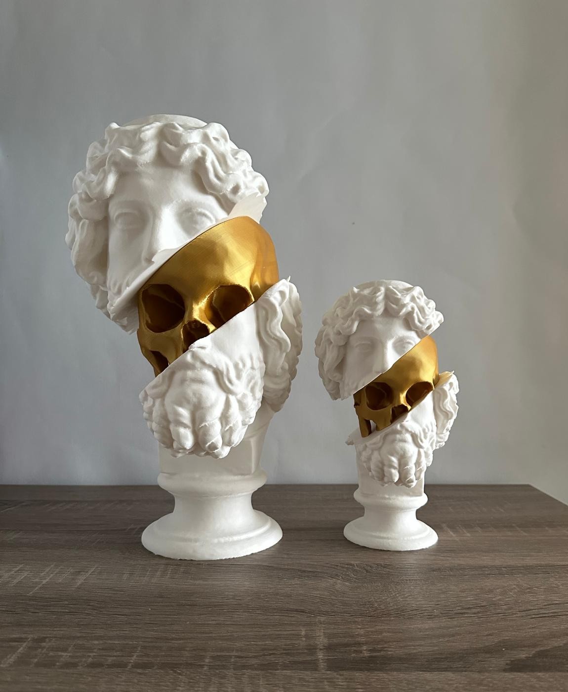 Dois bustos brancos com faixas douradas revelando crânios dourados, sobre mesa de madeira e fundo cinza.