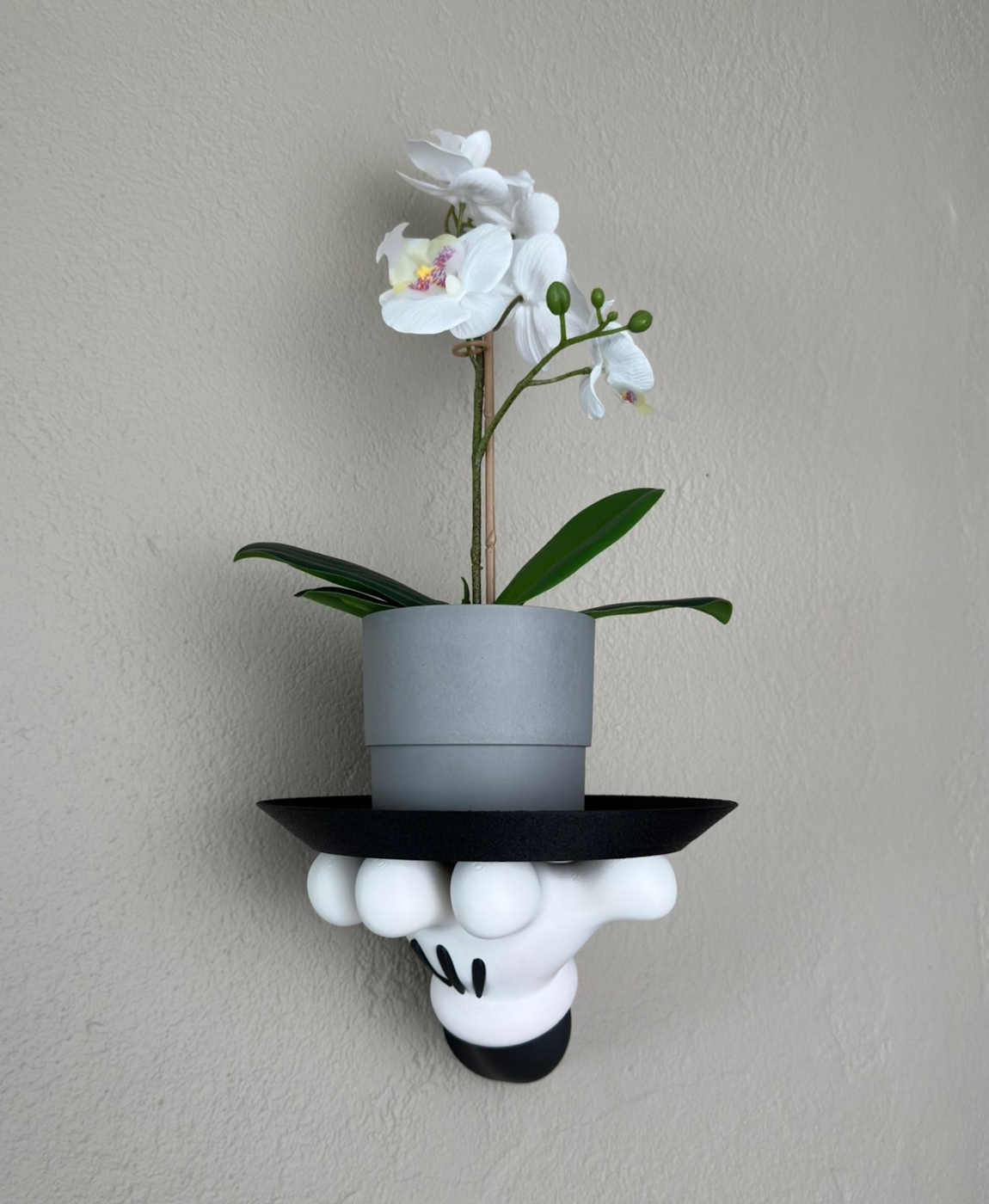 Orquídea branca em vaso cinza sobre suporte de parede decorativo em forma de mão com chapéu preto.